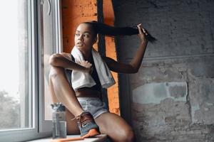 Attractive young African woman adjusting hair while sitting on the window sill in gym photo