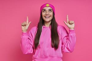 Beautiful young woman in funky hat pointing up and smiling against pink background photo