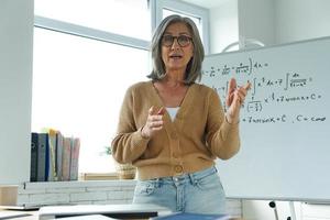 anciana enseñando matemáticas mientras está de pie cerca de la pizarra y gesticulando foto