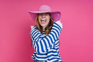 Excited young woman in pink hat hugging self while standing against colored background photo