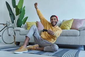 Excited young African man playing video games and gesturing while sitting on the floor at home photo