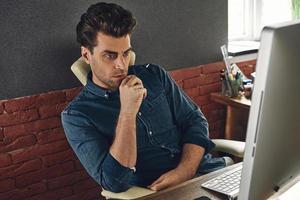 Thoughtful young man looking at the computer while sitting at his working place in office photo