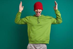 Happy young man pulling his hat on face and smiling while standing against green background photo