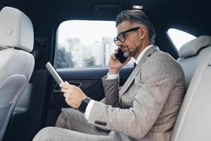 hombre de negocios maduro confiado que usa una tableta digital y habla por teléfono mientras está sentado en el auto foto