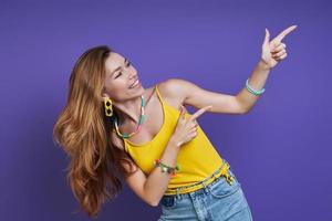 Attractive young woman pointing away and smiling while standing against purple background photo