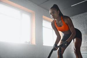 Concentrated young African woman preparing for training with battle ropes in gym photo