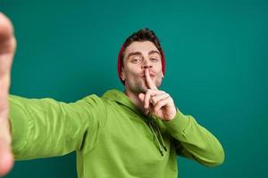 Handsome man making selfie and holding finger on lips while standing against green background photo