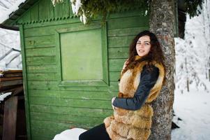 Elegance curly girl in fur coat at snowy forest park agasinst green wooden house at winter. photo