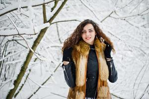 Elegance curly girl in fur coat at snowy forest park at winter. photo