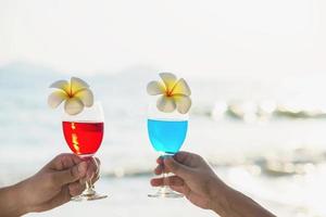 Pareja asiática sosteniendo una decoración de copa de cóctel con flor de plumeria con fondo de playa de mar de olas - feliz celebración de vacaciones de relax en el concepto de naturaleza marina foto