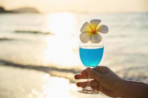 Closeup woman hand holding cocktail glass decoration with plumeria flower with beach background - happy relax celebration vacation in sea nature concept photo