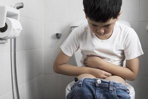 niño asiático sentado en la taza del inodoro sosteniendo papel tisú - concepto de problema de salud foto