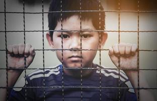 Dark portrait of a boy stand behind and holding steel screen or chain link fence - stressed sad child with no freedom concept photo
