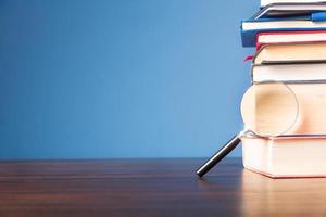 Stack book with magnifying glass on wooden desk in information library of school or university, concept for education, reading, study, copy space on blue background. photo