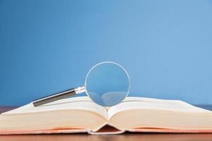 open book with magnifying glass on wooden desk in information library of school or university, concept for education,reading , study, copy space and blue background. photo