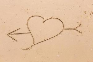 corazón y flecha dibujados en la suave arena de la playa. concepto de amor foto