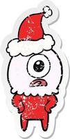 distressed sticker cartoon of a cyclops alien spaceman wearing santa hat vector