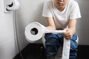 Asian boy sitting on toilet bowl holding tissue paper  - health problem concept photo