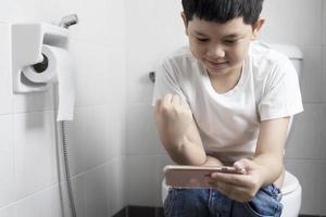 Asian boy sitting on toilet bowl while playing mobile phone - health problem concept photo