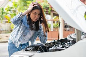 Angry Asian woman and using mobile phone calling for assistance after a car breakdown on street. Concept of vehicle engine problem or accident and emergency help from Professional mechanic photo