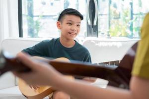 un niño asiático tocando la guitarra con su padre en la sala de estar por enseñarle a su hijo a tocar la guitarra, sentirse apreciado y alentado. concepto de una familia feliz, aprendizaje y estilo de vida divertido, amar los lazos familiares foto