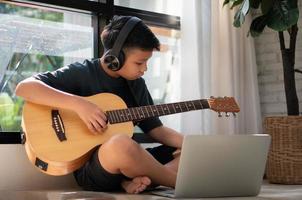 Asian boy playing guitar and watching online course on laptop while practicing for learning music or musical instrument online at home. Boy students study online with video call teachers play guitar. photo