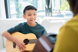 Asian boy playing guitar with father in the living room for teaching him son play guitar, feel appreciated and encouraged. Concept of a happy family, learning and fun lifestyle, love family ties photo