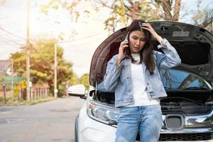 mujer asiática enojada y usando un teléfono móvil pidiendo ayuda después de una avería en el coche en la calle. concepto de problema del motor del vehículo o accidente y ayuda de emergencia de un mecánico profesional foto