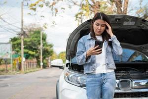 mujer asiática enojada y usando un teléfono móvil pidiendo ayuda después de una avería en el coche en la calle. concepto de problema del motor del vehículo o accidente y ayuda de emergencia de un mecánico profesional foto