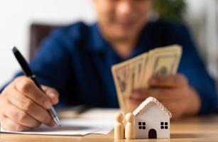 Selective focus for House model and figures people in front Happy Asian man holding a banknote and do financial accounting, Concept for financial planning, mortgage estate, Insurance, and home loan photo