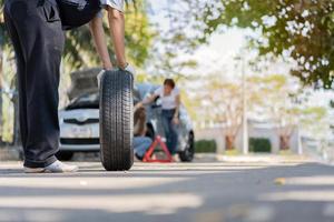 Expertise mechanic man in uniform holding a tire for help a woman for changing car wheel on the highway, car service, repair, maintenance concept. photo