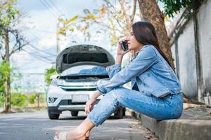 mujer asiática enojada y usando un teléfono móvil pidiendo ayuda después de una avería en el coche en la calle. concepto de problema del motor del vehículo o accidente y ayuda de emergencia de un mecánico profesional foto