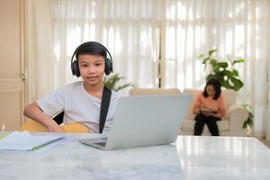 Asian boy playing guitar and watching online course on laptop while practicing for learning music or musical instrument online at home. Boy students study online with video call teachers play guitar. photo