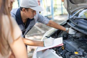 Asian male auto mechanic holds a clipboard and examines car engine breakdown problem with women customer and explain the root cause and estimate repair quotation, Car repair, and maintenance concept. photo