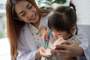 Asian female dentist teaching cute little girl brushing teeth with toothbrush and stomatologist telling girl child about oral hygiene in dental clinic, Education and prevention cavities concept. photo