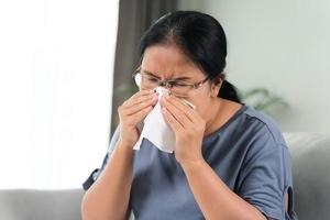 una mujer enferma de frío tiene tos alérgica a la nariz o estornuda con papel tisú sentado en el sofá. concepto médico y de salud.
