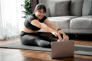 una mujer gordita asiática sentada en el suelo en la sala de estar practica una lección de yoga en línea con la computadora. mujer con clase de entrenamiento de meditación en la computadora portátil. foto
