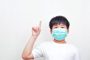 el niño asiático con una máscara quirúrgica médica, con una camiseta blanca y apuntando hacia arriba. foto