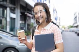 Young student Asian woman holding coffee and tablet wearing face shield to prevent virus photo