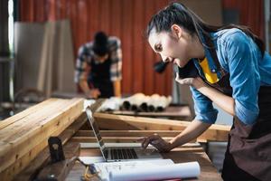 una joven carpintera asiática que usa una computadora portátil mientras habla con un cliente por teléfono en un taller de carpintería de madera. foto