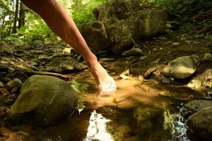 Elegant move of a barefoot woman, with a closeup of taking her foot out of a clean little puddle of clear fresh water, in the countryside surrounded by rocks and pebbles, in the shades of trees photo