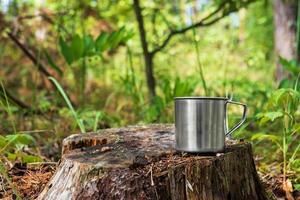 Metal tourist mug with hot drink stands on stump in the forest. photo