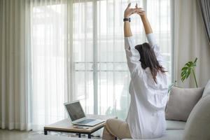 joven mujer de negocios con pantalones blanco sentada en un sofá con una laptop levantando los brazos por encima de la cabeza estirándose para aliviar el dolor después de estar sentada durante mucho tiempo en el trabajo. foto