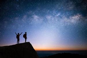 silueta de dos jóvenes de pie, con los brazos abiertos y mirando la estrella, la vía láctea y el cielo nocturno en la cima de la montaña. Disfrutaron viajar y tuvo éxito cuando llegó a la cima. foto