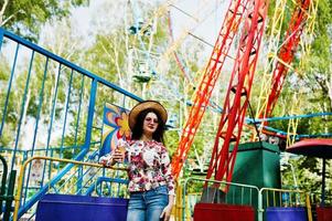 retrato de una chica morena con gafas rosas y sombrero con helado en el parque de atracciones. foto