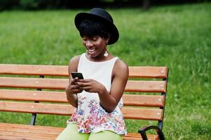 Increíble mujer modelo afroamericana con pantalones verdes y sombrero negro posada en un banco en el parque y enviando mensajes de texto por teléfono móvil. foto