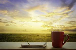 a red cup of coffee and note pad on wooden table and Empty grassland and sky at evening time photo