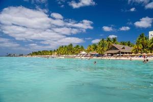 People swimming near white sand beach with umbrellas, bungalow bar and cocos palms, turquoise caribbean sea, Isla Mujeres island, Caribbean Sea, Cancun, Yucatan, Mexico photo