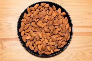 Whole almond nuts in black plate on the wooden background. Healthy vegetarian snack. Close-up photo. photo