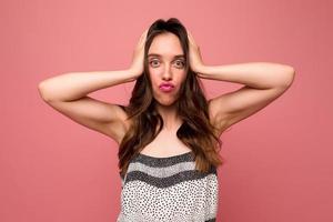 Funny adorable lady with wavy brown hair with pink lips is making faces and touching head over pink background. photo